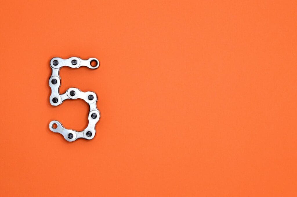 Creative arrangement of a chrome bicycle chain forming the number 5 on a vibrant orange background.